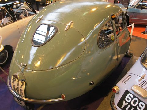 Grey-green rounded tail with an oval rear window and British plate, other microcars pressed close around it. Opens a larger view