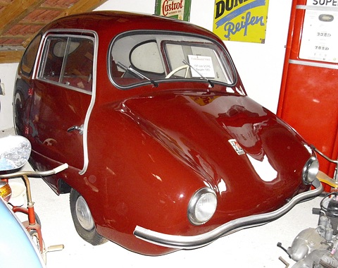 Dark red rounded three-wheeler with a curved chrome bumper in front of old Castrol and tyre signs. Opens a larger view