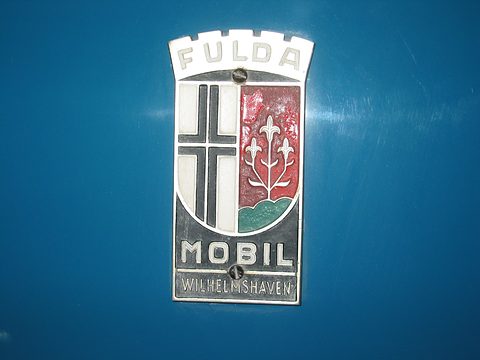 Enamel shield badge with a black cross and red panel of lilies, lettered FULDA, MOBIL and WILHELMSHAVEN, on blue paint. Opens a larger view