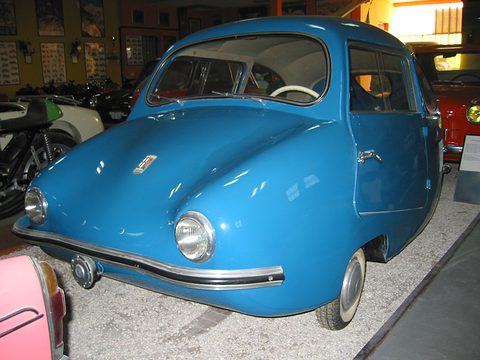 Blue rounded three-wheeler with chrome bumper and whitewall tyres on a gravel museum display. Opens a larger view