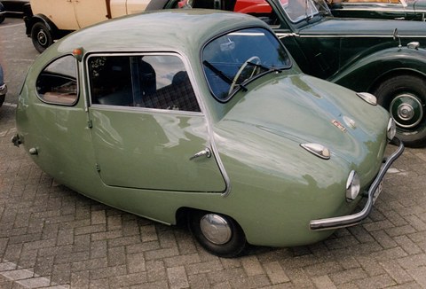 Grey-green rounded three-wheeler with tubular front bumper parked on brick paving beside a dark saloon. Opens a larger view