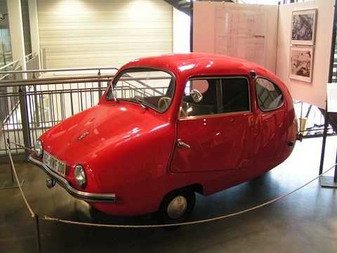 Glossy red egg-shaped three-wheeler with chrome bumpers behind a low rope in a bright museum hall.