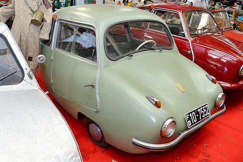 Pale green rounded three-wheeler on a red carpet between a hammered-aluminium car and a red saloon. Opens a larger view