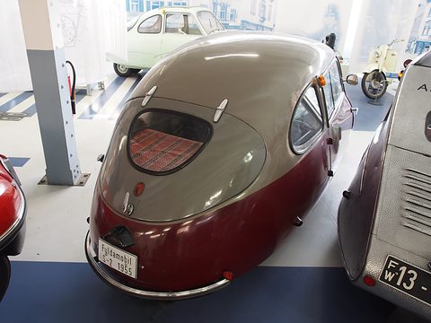 Grey-roofed rounded tail with an oval rear window and a display plate, a scooter and a white car behind. Opens a larger view