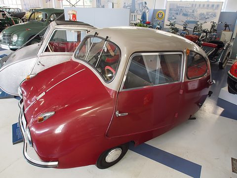 Red and grey three-wheeler in side view, its front-hinged door open, a hammered-aluminium Fuldamobil beside it. Opens a larger view