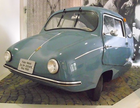 Blue-grey rounded car with chrome strips on a cobbled plinth, black-and-white period photographs behind. Opens a larger view