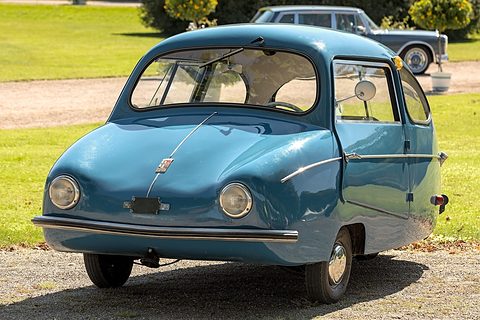 Blue rounded car parked on gravel by a lawn in sunshine, a black limousine behind. Opens a larger view