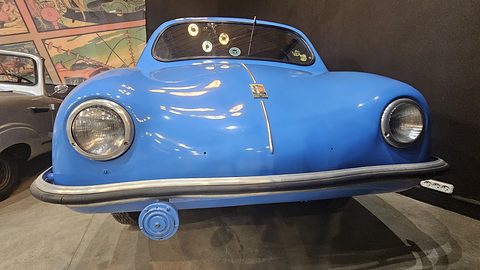 Blue rounded car facing the camera, crest badge on the bonnet, a colourful mural behind. Opens a larger view
