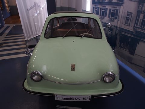 Pale green rounded car facing the camera with a “Fuldamobil S-7 1961” plate, a period street photograph behind. Opens a larger view