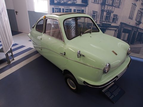 Pale green three-wheeler with a fabric sunroof on a blue-striped floor before a large period street photograph.