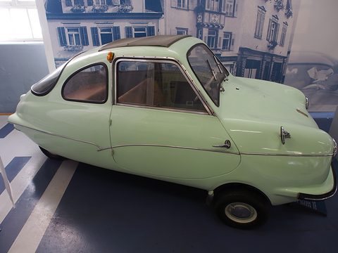 Side view of a pale green finned three-wheeler on a dark plinth before a printed street scene. Opens a larger view