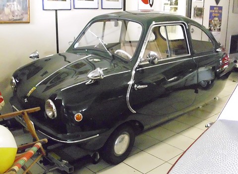 Dark green rounded car with chrome bullet mirrors and door handles on a tiled museum floor among beach props. Opens a larger view