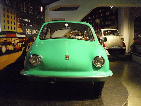 Turquoise rounded car facing the camera on a dark plinth, a painted city-night backdrop behind. Opens a larger view