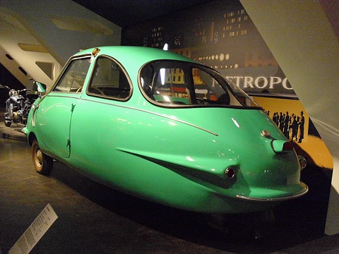 Turquoise three-wheeler with tail fins and chrome flank strips on a dark plinth before a mural. Opens a larger view