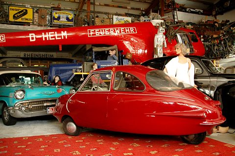 Red finned three-wheeler on a red carpet beside a turquoise American car, mannequins and a fire-brigade crane behind. Opens a larger view