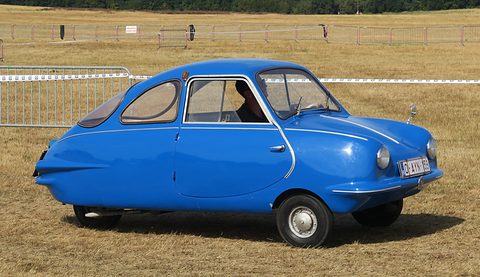 Bright blue finned three-wheeler on a dry grass airfield, crowd barriers and a tree line behind. Opens a larger view