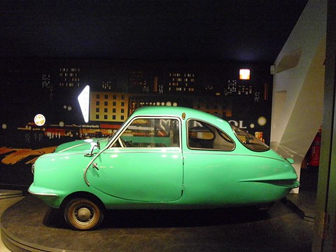 Turquoise three-wheeler in side view with a single front door and finned tail, against a dark painted backdrop. Opens a larger view