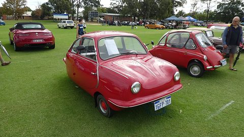 Red rounded three-wheeler on a sports field, a red Heinkel bubble car and other cars behind. Opens a larger view