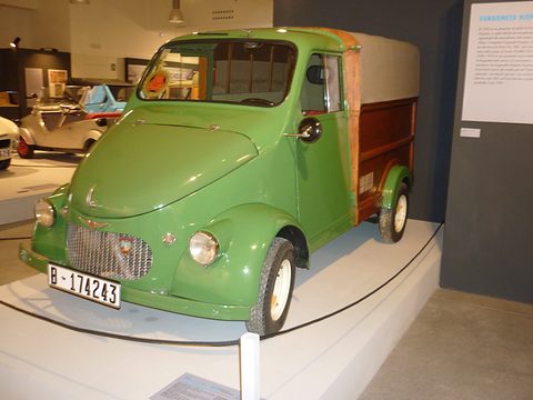Green small van with a mesh grille and a varnished wooden box body with a canvas top, on a museum plinth. Opens a larger view