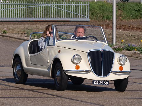 A small white open roadster with a tall pointed chrome grille and round headlamps, a man and a woman aboard. Opens a larger view