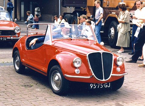 An orange open roadster with a tall black and chrome grille on a brick street, onlookers and a lorry behind. Opens a larger view