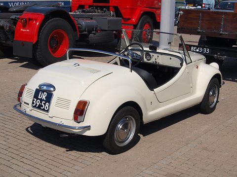 Cream open two-seat roadster from the rear left beside a red and black lorry, on brick paving.
