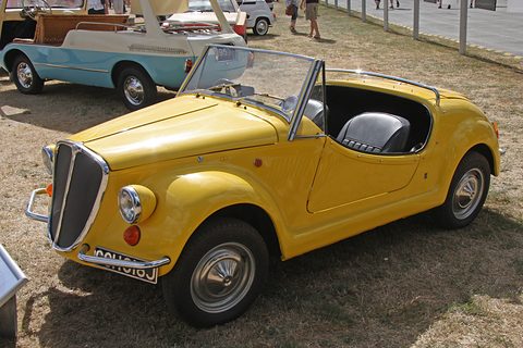 Yellow open roadster with a tall chrome-framed vertical grille and separate front wings, parked on dry grass. Opens a larger view