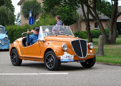 An orange open roadster with a tall chrome grille and black alloy wheels on a village road, two people aboard. Opens a larger view