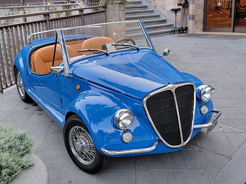 Blue open roadster with a tall black and chrome grille, wire wheels and tan seats, seen from the front right.