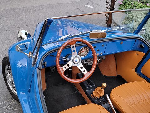 Looking down into a blue open roadster with tan leather seats, a wooden three-spoke steering wheel and a round speedometer. Opens a larger view