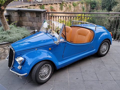 Blue open roadster with tan seats, wire wheels and flowing separate wings, parked on stone paving above a gorge. Opens a larger view