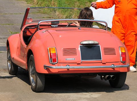 Rear of a coral-pink open roadster with twin exhausts and louvred engine lid, a marshal in orange overalls beside it. Opens a larger view