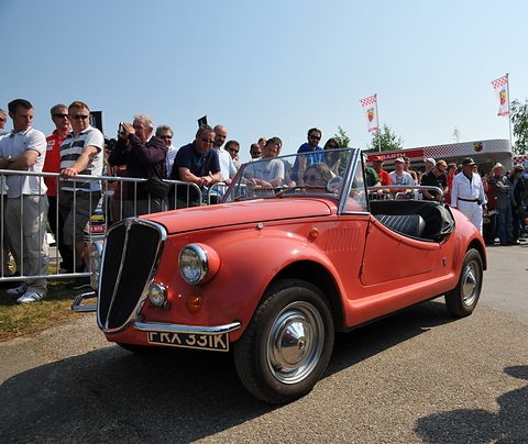 Red open roadster with an upright chrome grille in front of spectators lined up along a barrier on a sunny day. Opens a larger view