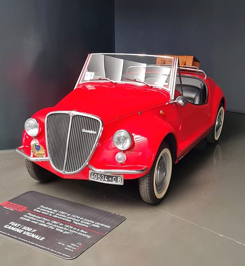 Red open roadster with whitewall tyres and a vertical chrome grille on a grey plinth, an information plaque before it.