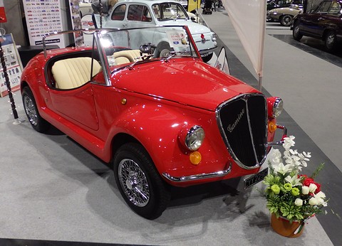 Red open roadster with cream seats, wire wheels and Gamine script on its grille, a vase of flowers by its nose. Opens a larger view