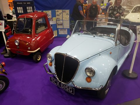 Pale blue open roadster next to a tiny red one-seat car on purple carpet, a blue police-box prop behind.
