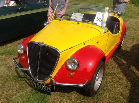 Yellow open roadster with red wings and an upright chrome grille, a show card on the windscreen, on grass. Opens a larger view