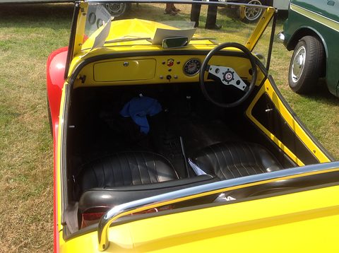 Looking down into a yellow open roadster with two black ribbed seats, a three-spoke wheel and a yellow painted dash. Opens a larger view