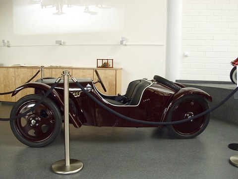 Low maroon open two-seater with disc wheels and swept wings, seen from the left side behind a rope barrier.