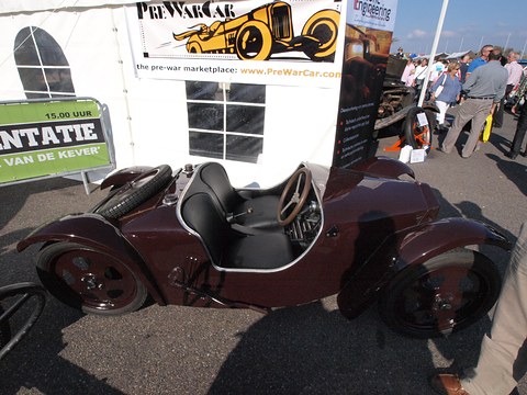 Low maroon open two-seater with black seats parked outdoors in front of a marquee and event banners. Opens a larger view