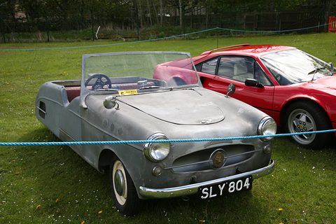 Grey open three-wheeler with a chrome grille and bumper on grass behind a blue rope, a red sports car beside it.