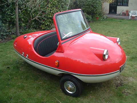 Red and cream bubble-shaped three-wheeler with a black seat on a lawn before shrubs. Opens a larger view