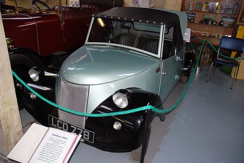 Pale green open two-seater with a black hood and mesh grille behind a green rope, a label board in front.