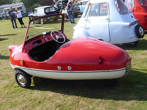 Red and cream bubble-shaped open three-wheeler on grass, a pale blue Isetta beside it. Opens a larger view