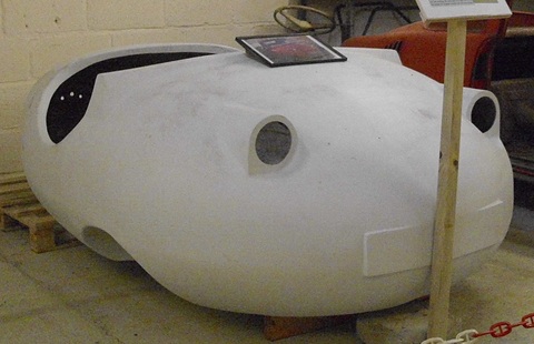 Side view of a grey glass-fibre bubble car shell with an oval opening, propped on pallets. Opens a larger view