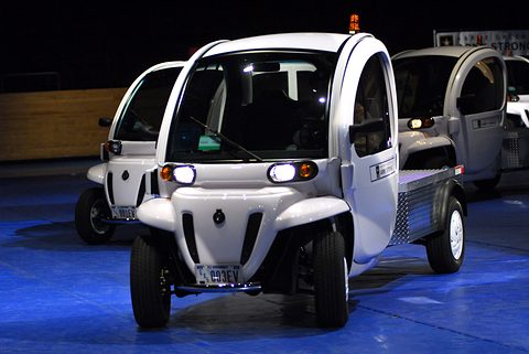 White two-seat electric utility vehicles with headlamps lit lined up on a blue floor in a dark hall. Opens a larger view