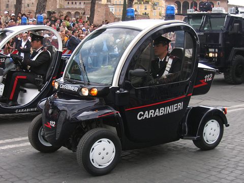 Dark blue electric car lettered CARABINIERI with a blue roof light and a uniformed driver, another behind. Opens a larger view