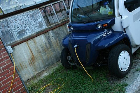 Blue nose of a small electric car with a yellow cable plugged into a socket on a brick wall, grass below. Opens a larger view