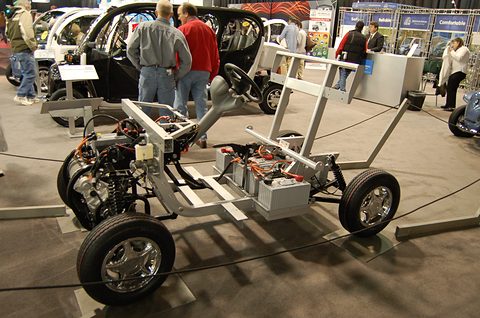Bare chassis with a row of batteries, wheels, steering column and seat frame on a show floor.