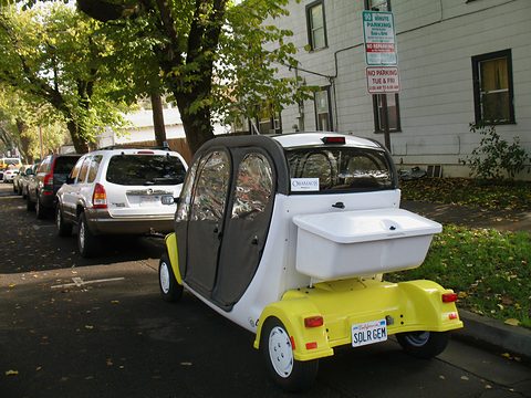 Rear of a white two-seat electric car with a yellow lower body, a cargo box and a SOLR GEM plate at a kerb. Opens a larger view